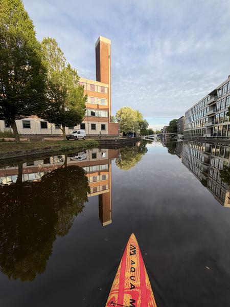 The photo captures a serene view from a paddleboard on a canal in Sarphatistraat, Weesperbuurt, Amsterdam, reflecting the surrounding modern buildings and lush green trees